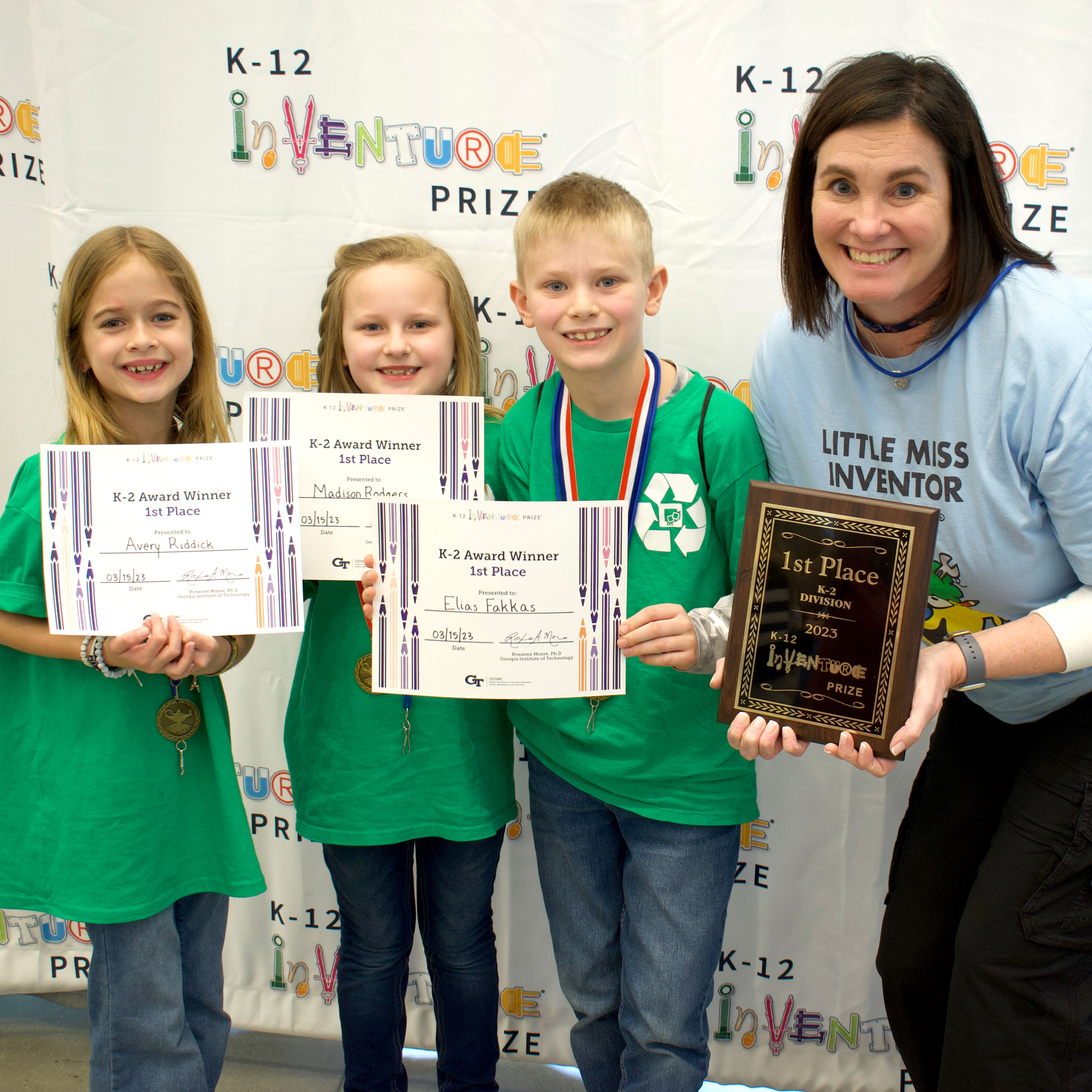 Three smiling students holding up certificates and standing with a smiling teacher holding a plaque