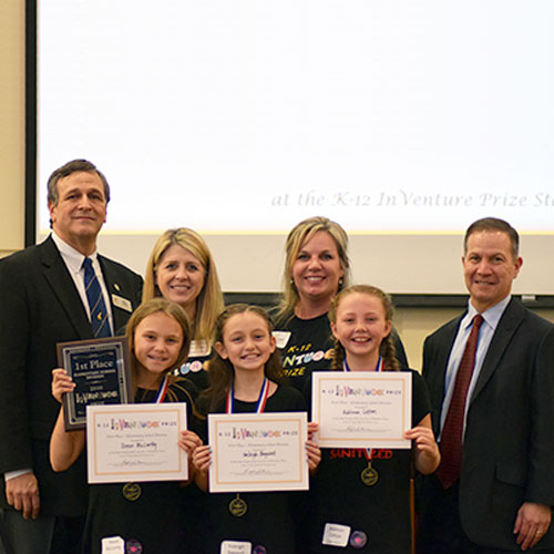 Three smiling students holding up certificates and a plaque in front of four smiling adults