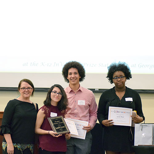 Three smiling students holding up certificates and a plaque and standing beside a smiling adult