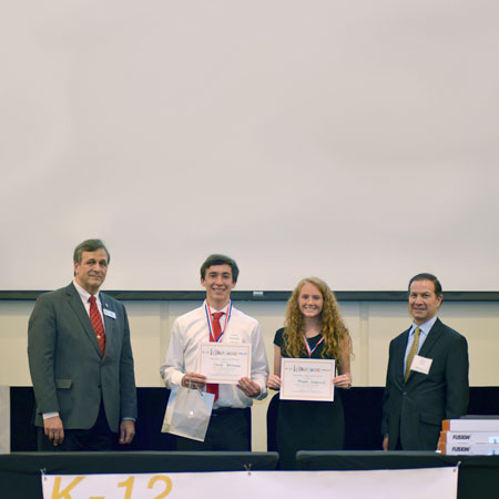 Two smiling students holding up certificates and standing beside two smiling adults