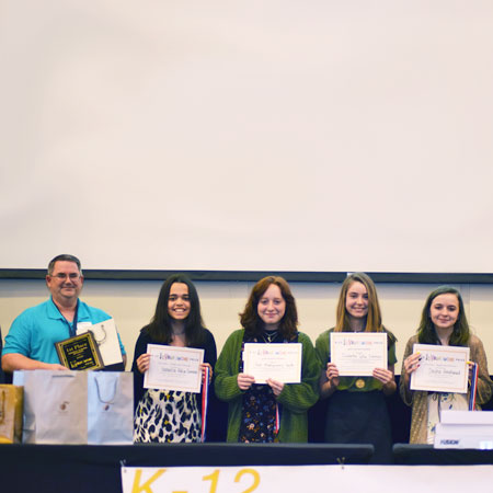 Four smiling students holding up certificates and standing beside a smiling teacher holding up a plaque