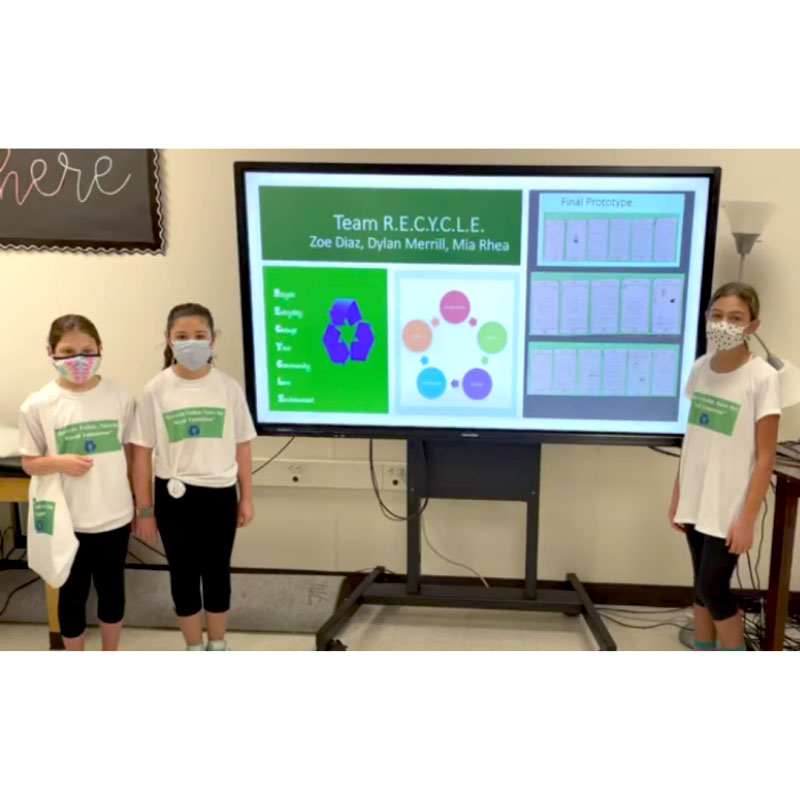 Three students in face masks in front of a screen showing their project