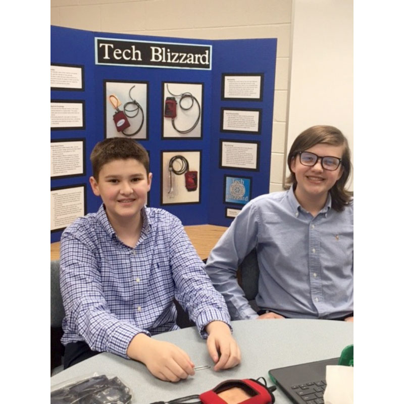 Two smiling students sitting at a table with a triboard of their project behind them