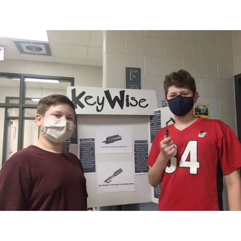 Two students in masks in front of their project triboard with one of the students holding up their prototype