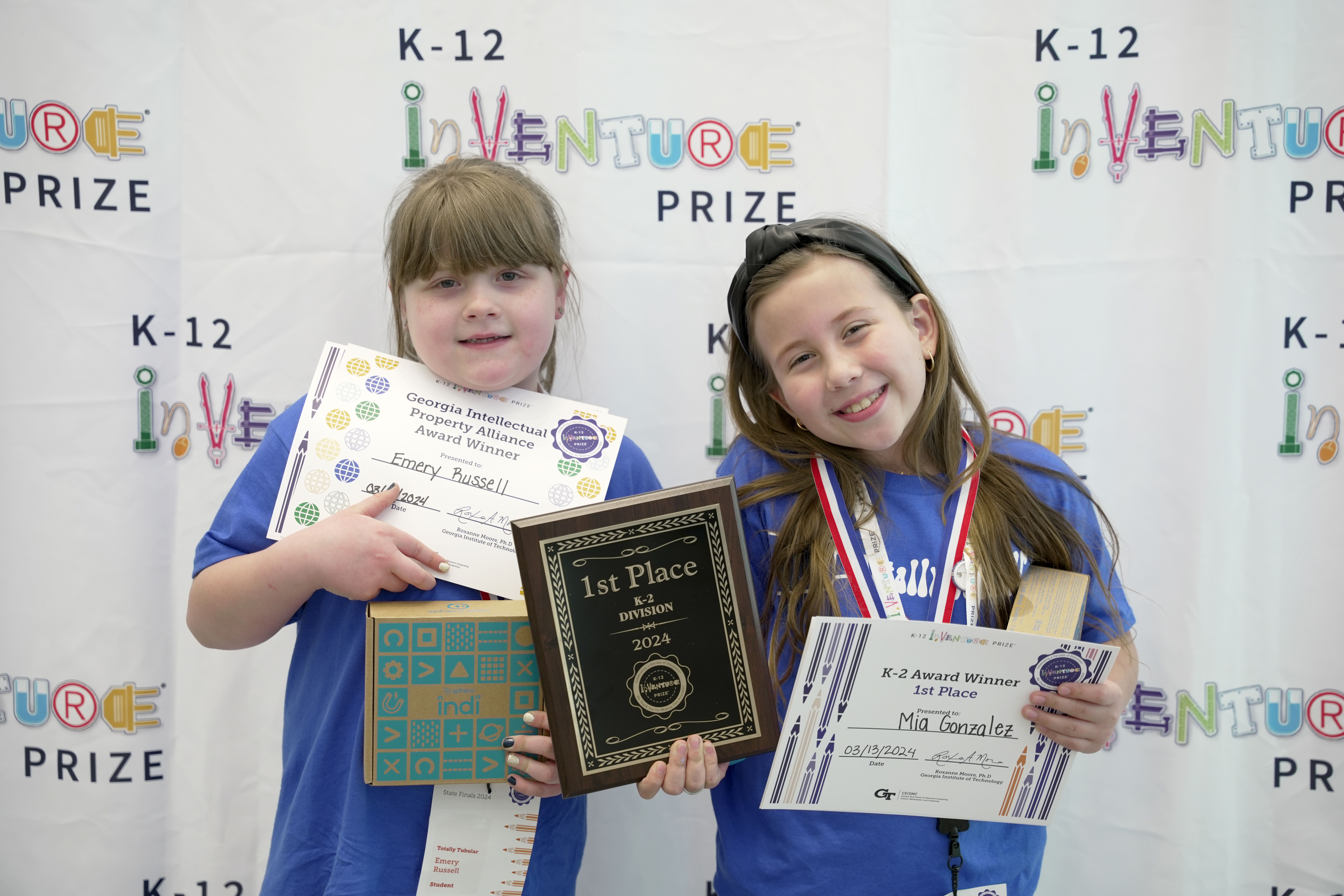 Two smiling students holding certificates, prizes, and a plaque