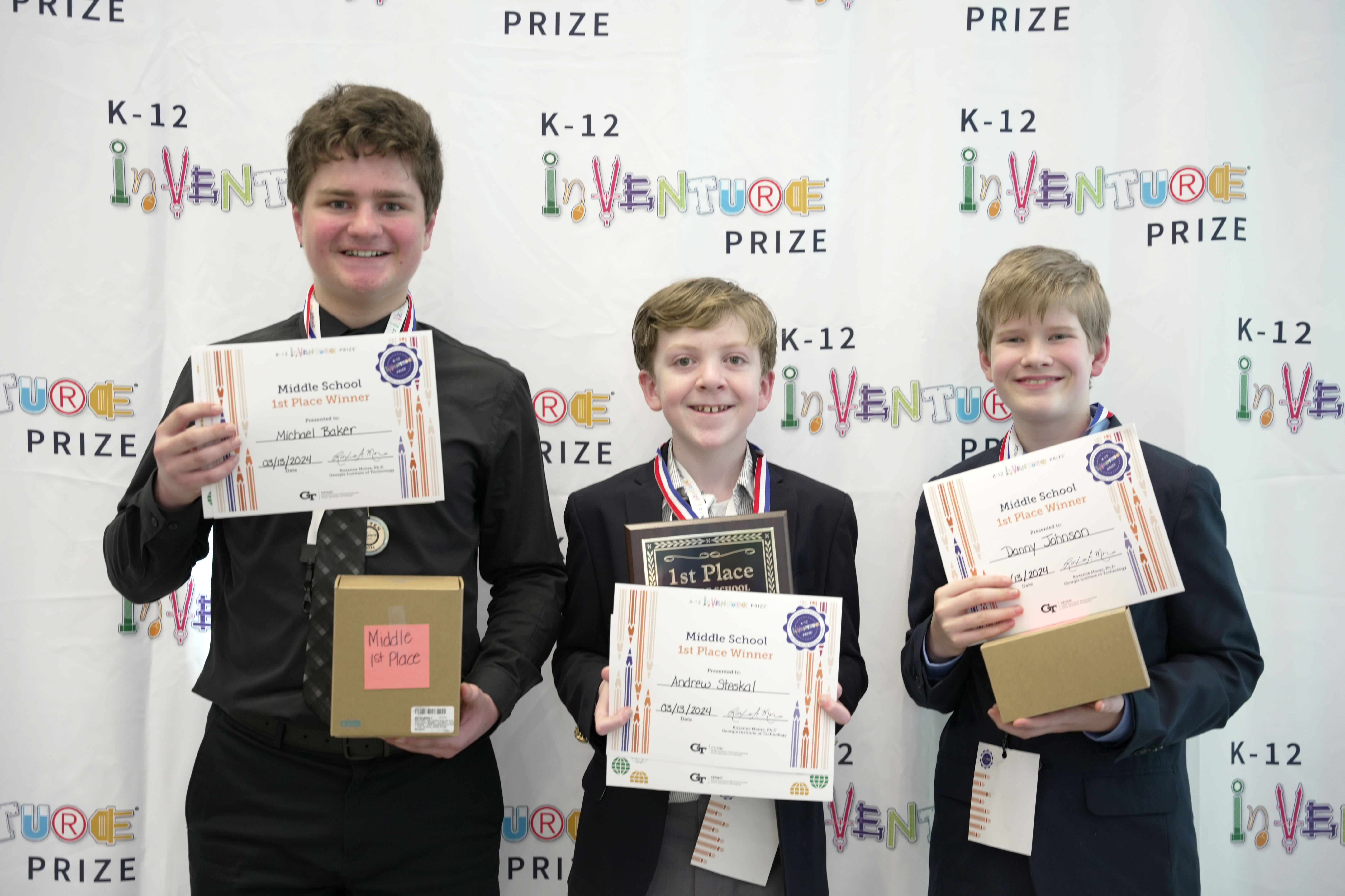 Three smiling students holding certificates, prizes, and a plaque