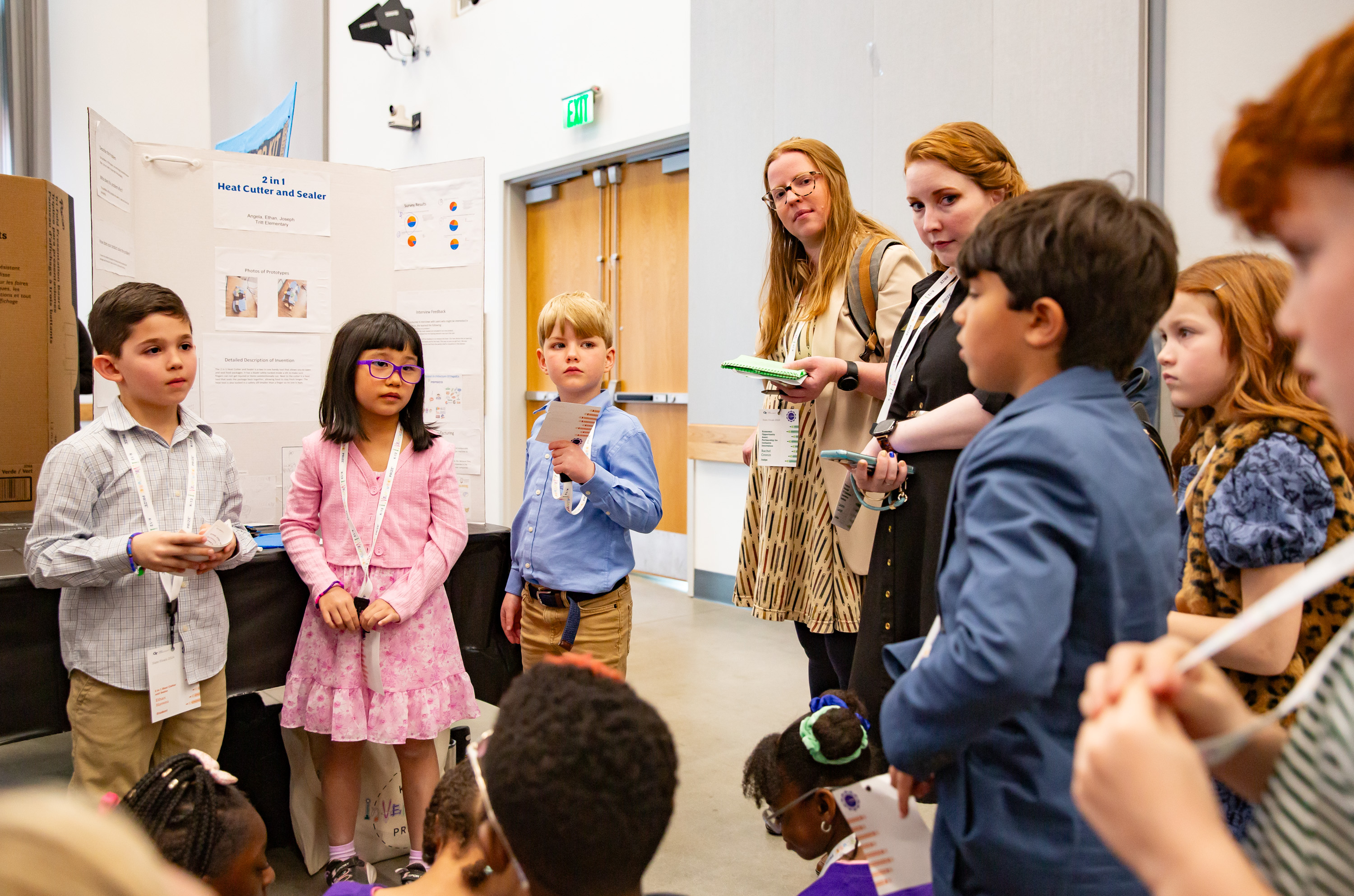 A group of students asking another group of students questions with two adults in the back and another group of students sitting