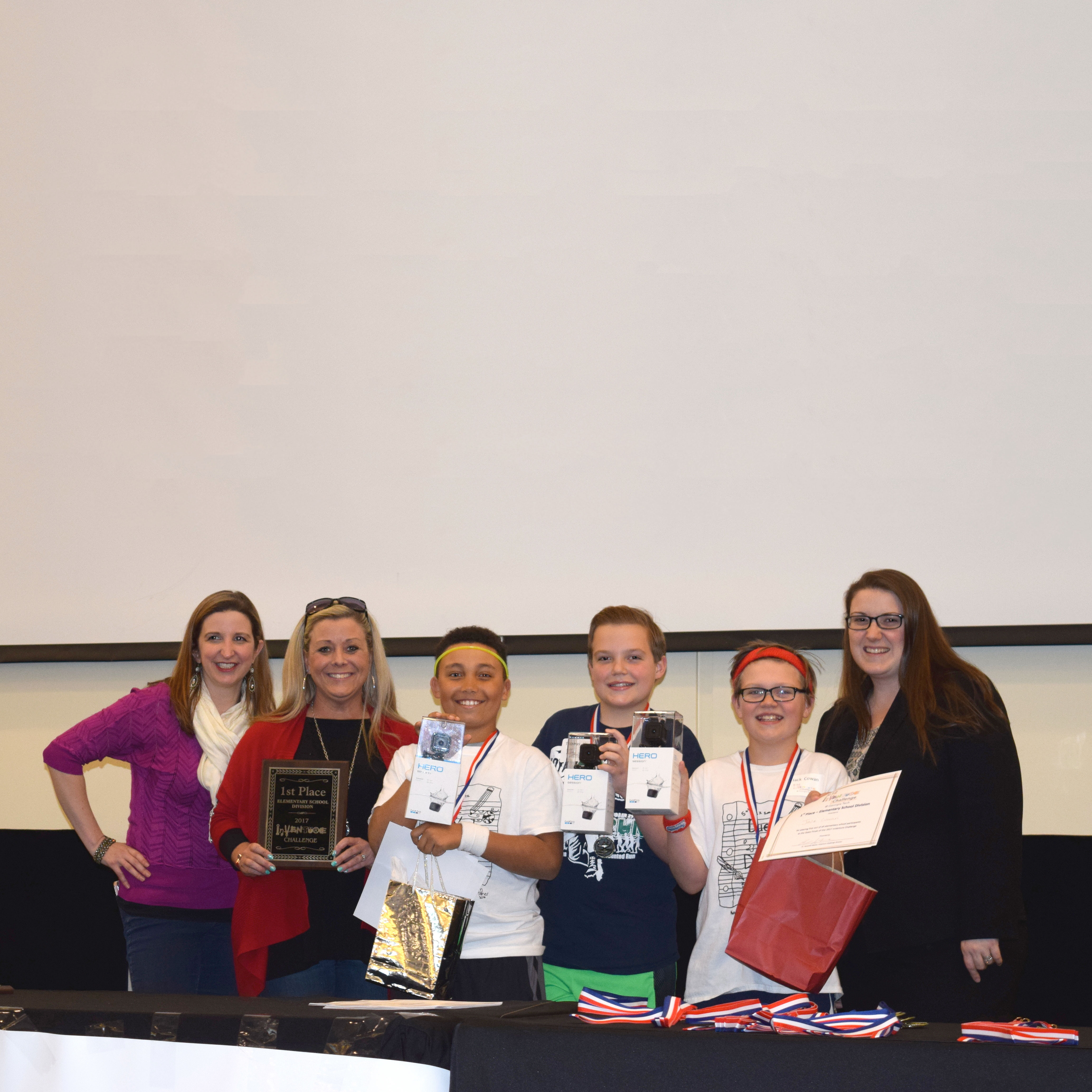 Three smiling students holding up certificates and prizes and standing next to three smiling adults