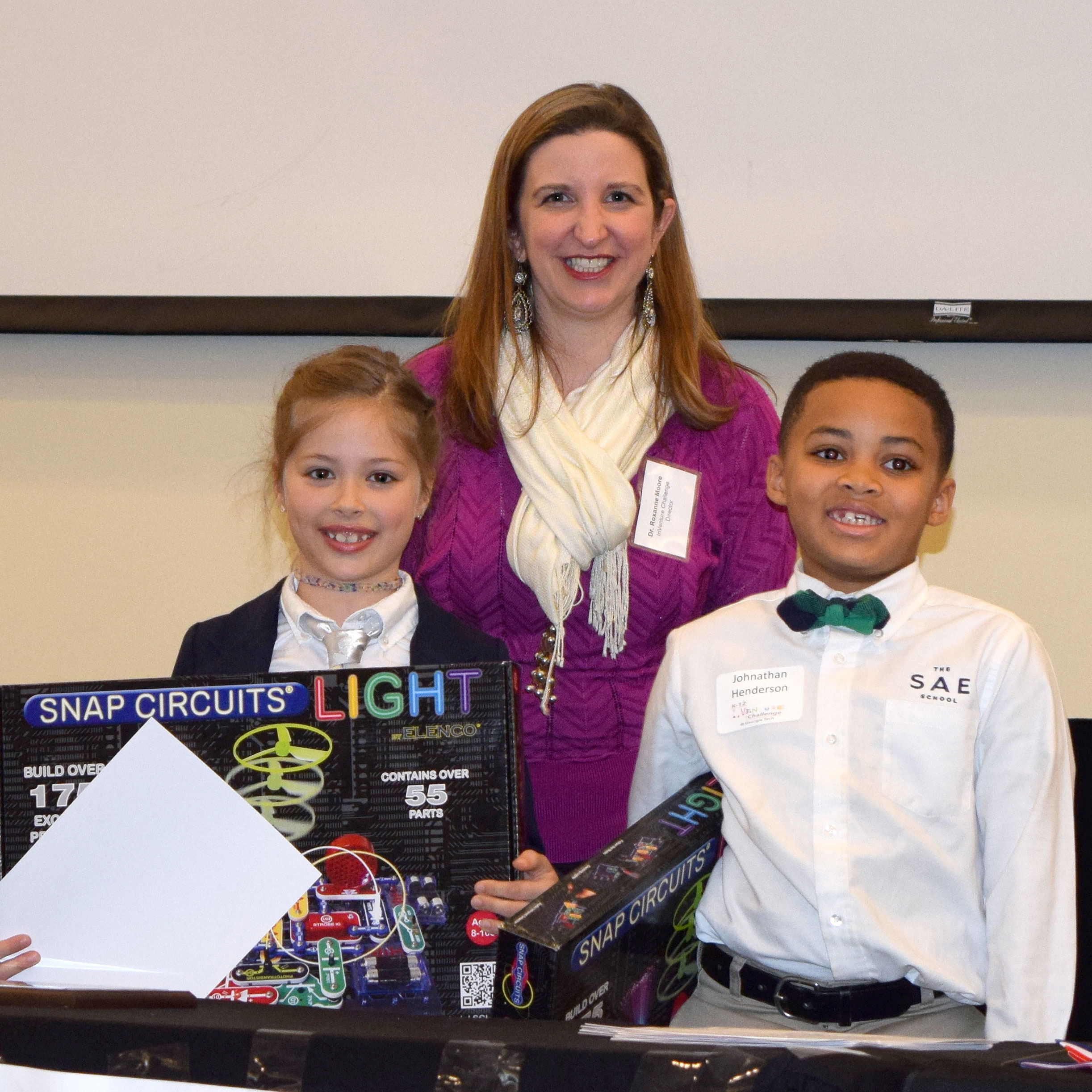 Two smiling students holding up prizes and standing in front of a smiling adult